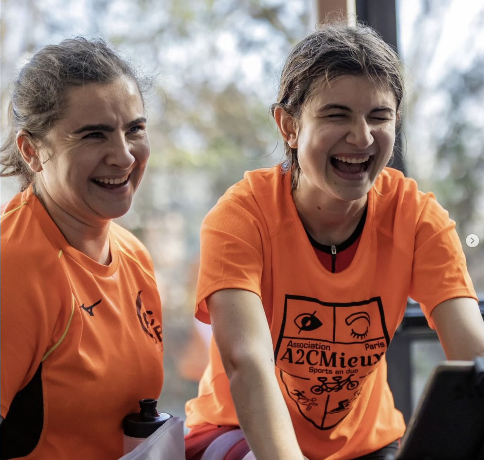 Deux sportives de l'association A2CMieux, en maillot orange, souriant pendant une séance de vélo.