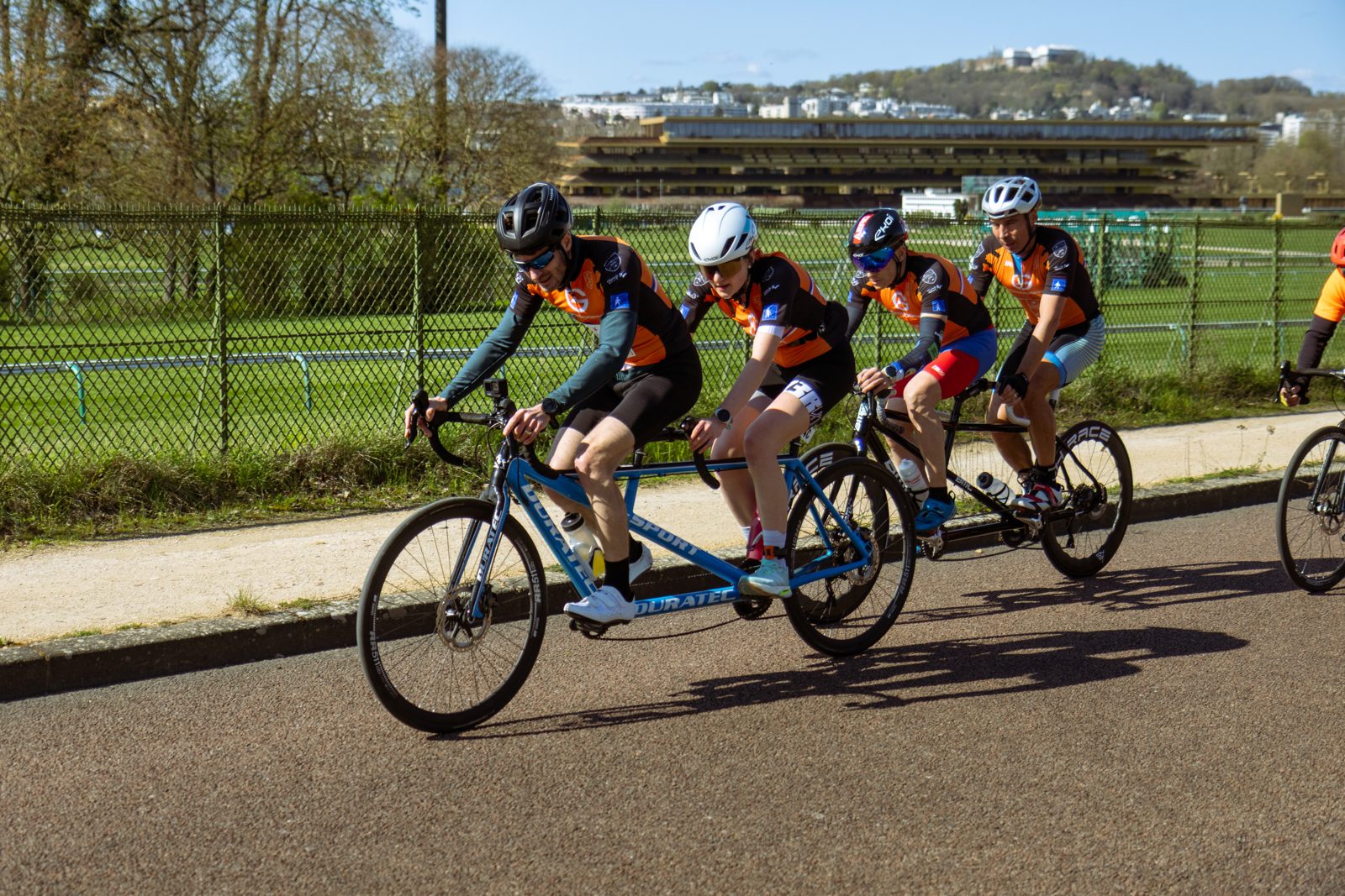 Deux tandems de l'association A2CMieux en pleine course sur route, pilotes et équipiers en maillot orange et noir.